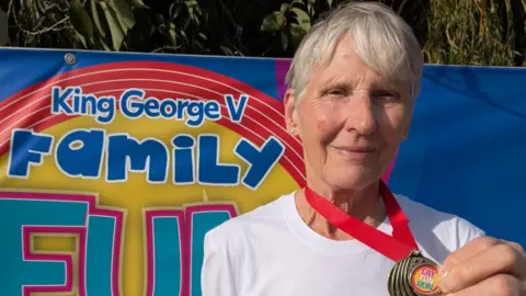 Lincs Inspire A woman with short grey hair is wearing a white top and holding up a medal which says Cry Fun Run on it. Behind her is a colourful banner which says King George V Family Fun.