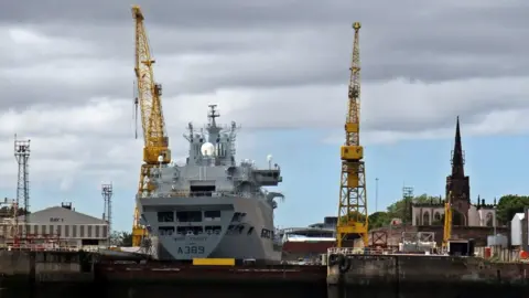 El Pollock/Geograph A ship is in the Cammell Laird shipyard. There are two yellow cranes and Birkenhead Priory is to the right.