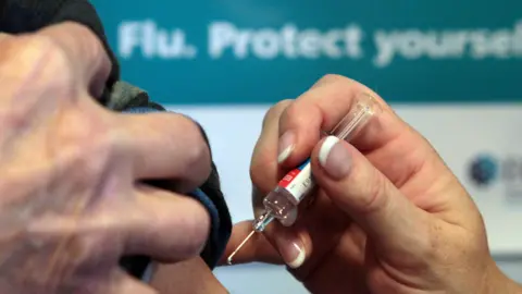PA Media Close up of a syringe with a flu vaccination, ready to go into someone's arm. The patient holds their sleeve up to get the medicine. A sign saying "Flu. Protect yourself" is blurred in the background.