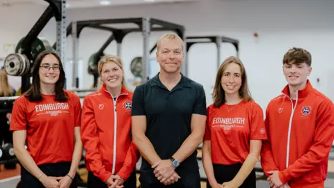 University of Edinburgh Three young women and a young man stand on either side of Sir Chris Hoy. Sir Chris is dressed in a black polo shirt and trousers. The students are wearing red Edinburgh University t-shirts and tracksuits. 