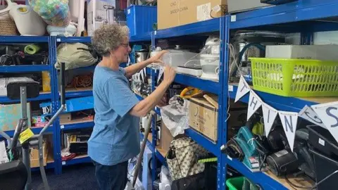 BBC A woman in a blue T-shirt stands against a blue rack of items including sanders, slow cookers and camping supplies.

She is arranging items on the rack.