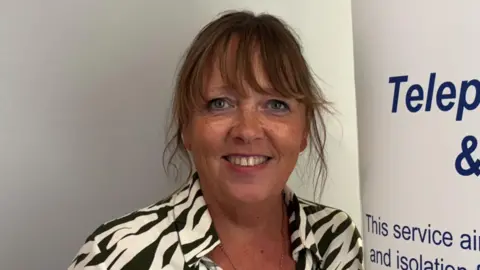 A woman with tied up brown hair is smiling and looking into the camera. she is wearing a black and cream open-neck pattern dress. 