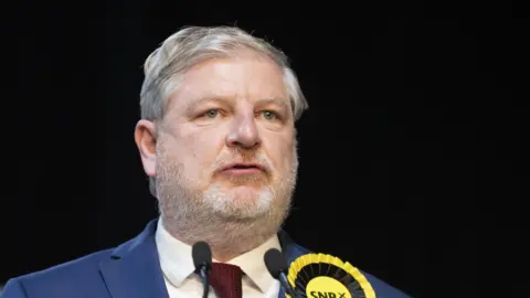 PA Media Culture secretary Anugs Robertson wears a blue suit, white shirt, burgundy knitted tie and yellow and black SNP rosette.  Two slim mics from a lectern can be seen at the bottom of the image. 