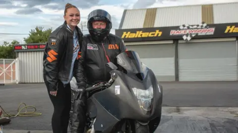 Danny Cockerill Danny's daughter stands next to him while he sits on his racing bike. He has the helmet on with visor up and wears a black jacket with the word DAN in orange. She wears a Santa Pod leather jacket too with the number 21 on a badge, over a blue t-shirt and black leggings. 
