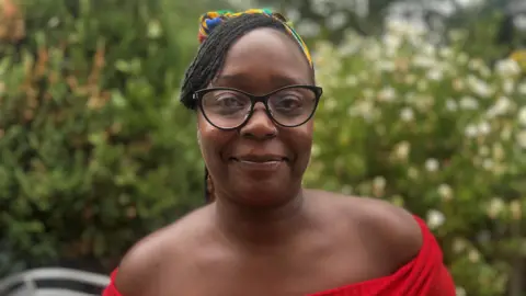 BBC Leasuwanna Griffith, who is wearing black rimmed glasses and a red strapless top and a colourful headscarf in her hair. She is smiling at the camera.