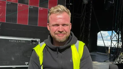 Shivani Chaudhari/BBC Chris Halls wearing a hi-vis jacket and a black hoodie. He is sitting on the main stage and smiling at the camera. He has blond hair and stubble.