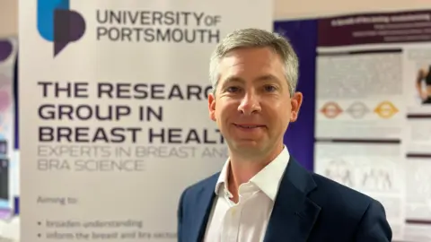 a man with short grey hair, wearing a white short and navy suit jacket, standing in front of a board with University of Portsmouth branding