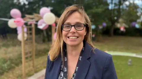 Heather Bide with black glasses and wearing a blue blazer smiles broadly at the camera with a garden decorated with pink balloons in the background