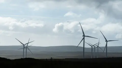 NurPhoto/Getty Images A view of a wind farm from Benbradagh Summit in County Londonderry on a cloudy day. There are seven white turbines with three spokes each in a field. There are rolling hills on the horizon.
