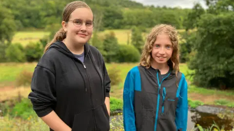 BBC Emily, on the left, with her brown hair in a ponytail. She is wearing a black zip up hoodie and glasses. Eva, right, wearing a blue and grey zip up jacket. She has blonde curly hair which is down. Both are looking at the camera and smiling. In the background is green grass and bushes.