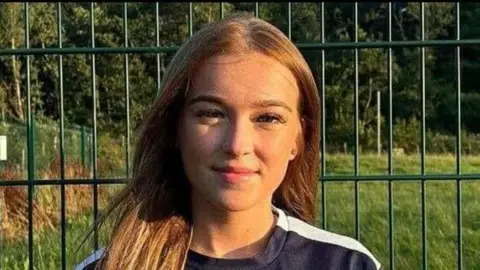 Charlene Paterson A girl with long brown hair smiles at the camera. She stands in front of a wire fence with grass behind it and she appears to be wearing a football strip
