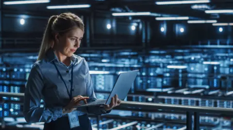 Getty Images A woman holding a laptop inside an AI data centre, which has lots of computer systems glowing blue in the background.