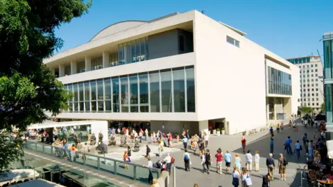 Getty Images An exterior photo of the Southbank Centre on a sunny day. There are people milling around outside the venue, a large concert venue with a glass frontage. 