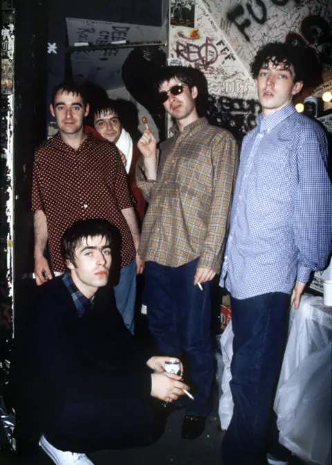 Dennis Morris Colour photograph of Oasis posing for the camera. All of them are stood up with the exception of Liam Gallagher who is crouched down.