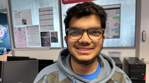 BBC An Asian man with glasses, smiling at the camera. He has a blue top on with a grey pull string jumper. His glasses are circular and large on his face, he's stood in front of a series of stacked up recycled PCs and looks excited to be talking on the matter 