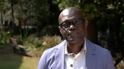 Gilbert is a middle aged black man. He is bald and wears a white shirt and a blue blazer with black glasses.