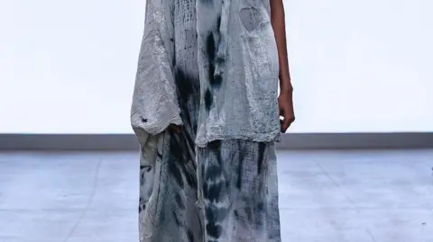 Gabrielle Thomas A close up of the lower portion of a design. A model is walking down the catwalk wearing a pale blue tattered sheet with darker blue stains and splashes.