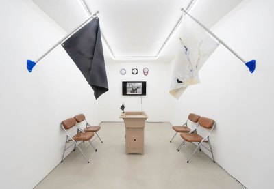 View of an installation in an art gallery, featuring foldable leather chairs facing a podium. Behind is a TV screen and three clocks. On the left and right walls is one large flag each.