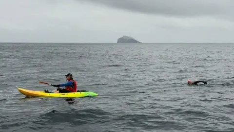 Dylan Antscherl A man is paddling on a canoe, followed by a woman in a full wet suit and cap, swimming behind him. They are in a vast expanse of water, with only Bass Rock visible on the horizon. 
