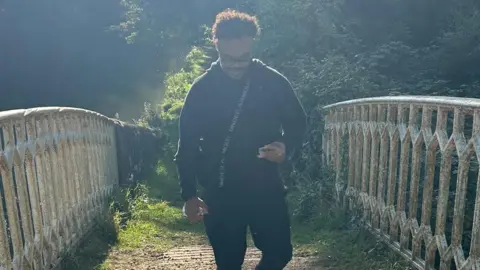 BBC A man wearing glasses, a black hoody and black trousers, with a strap across his front, walks across a bridge. Trees can be seen behind him.