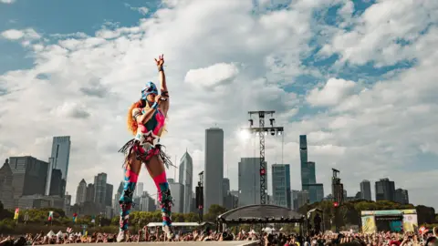 Josh Druding Chappell Roan wears a pink leotard emblazoned with stars, thigh-high boots with stars and a super hero mask. She is standing on a stage with one hand raised in the air.