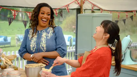 Channel 4/Love Productions Pui Man throws her head back in laughter while putting cake mixture into a silver bowl. She has her hair in a ponytail and is wearing a red jumper. In front of her is TV presenter Alison Hammond, who is wearing a blue dress and is also laughing.