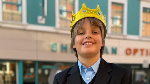 Rob Constantin/BBC Isaac is stood in the middle of the image looking towards the camera smiling. He has light brown hair and is wearing a gold crown on his head. He is wearing a blue shirt and a black blazer and is standing in front of a building which is painted blue.