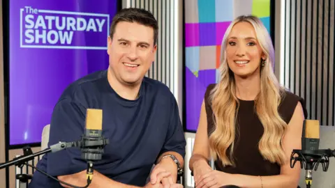 BBC A publicity shot of Steven Mill and Amy Irons in a BBC radio studios - Steven, with short dark hair and wearing a navy t-shirt sits in front of a mic beside blonde Amy, in a black sleeveless top, also with a microphone in front of her. A Saturday Show logo in white and purple iso a screen behind them.