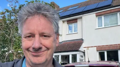 Michael De Podesta Michael with short hair stood in front of his house. He is smiling. There are solar panels on his roof and his house is cream with exposed brick.