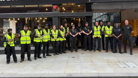 Belmont Guard Security Services A line of security guards in yellow hi-vis vests. Some are wearing black caps. There are seven on the left, then three police officers in uniform, followed by three security guards and two further men in black T-shirts and trousers. They are standing on light-coloured paving slabs in front of the glass frontages of two shops.