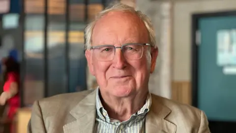 Tony Beckingsale is pictured wearing a cream jacket and a green and white striped shirt. Mr Beckingsale has white hair, is wearing glasses and is smiling. The background is blurred.