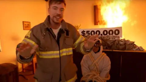 YouTube.com/@MrBeast MrBeast, dressed in a firefighters jacket is standing in a burning room in front of a man in a white hoody, who is tied up. Behind both of them is a table piled high with sacks of dollar bills.