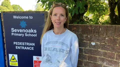 A woman in her early forties with long blonde hair smiles a the camera whilst wearing a grey sweatshirt. She stands next to a dark blue sign which reads : "Sevenoaks Primary School".