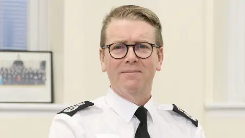 PA Media Chief Constable Will Kerr stands in front of a Devon and Cornwall Police sign. sits in an office. He is wearing a white shirt with a black tie and black epilates. A framed photo of police officers is behind him.