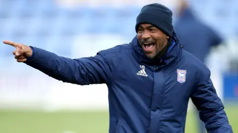 PA Media Kieron Dyer wearing a navy beanie hat and navy coat with the Ipswich Town and Adidas emblems on. He is laughing with his mouth open and standing on the pitch at a stadium, which has blue seats behind him. Dyer is pointing at something out of sight to his right. 