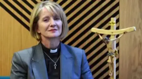 Church of England Bishop of Warrington Bev Mason with blond bobbed in front of wooden wall with black stripes on it and wooden figure of Jesus Christ on the cross.