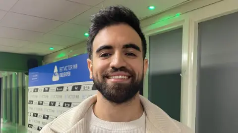 James Patient/BBC Faisal Labban has dark hair and a beard. He is smiling in front of windows with white panels and is wearing a cream jacket over a white T-shirt. Behind him is a white and blue board that has the tournament's sponsors on it.