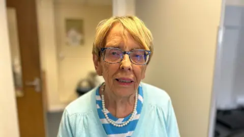 BBC A woman with short light coloured hair is looking into the camera. She is wearing dark rimmed glasses, a blue and white striped top and a light blue cardigan with a pearl necklace. 