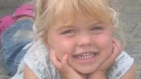Angie Bunce-Mason Elana is in the middle looking towards the camera smiling. She is laying on a concrete floor with her hands on her chin. She has blonde hair and a fringe.