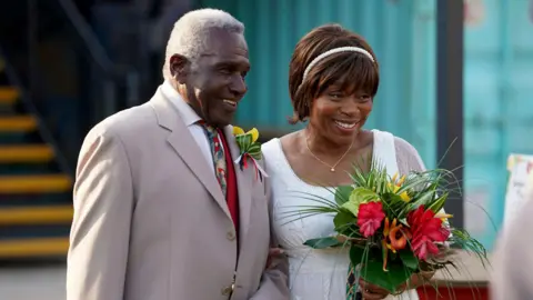 Kieron McCarron/BBC Rudolph Walker, in character as Patrick Trueman, is in a beige suit, white shirt and colourful tie. He is arm-in-arm with Angela Wynter, in character as Yolande Trueman, who is in a white dress. She is holding a bouquet of flowers.