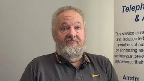 A man with grey hair and a grey beard looks towards the camera. He is wering a dark grey polo shirt with yellow details. 