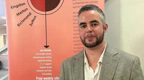 Joel Robinson, who has greying hair, a beard and dark eyebrows, is standing in front of a sign that provides information about the charity's outreach clinics in London. He is wearing a light-coloured shirt and a grey suit jacket and is looking at the camera.