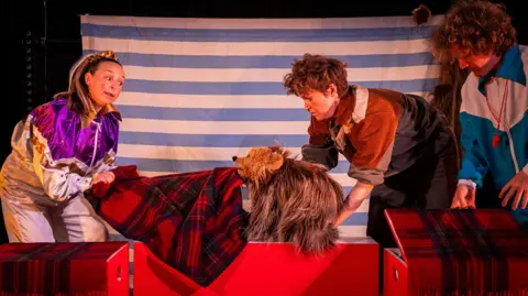 Eoin Carey A theatrical scene featuring three performers interacting with a bear-like puppet. The stage is decorated with red plaid-covered platforms and a striped blue-and-white backdrop. The performers wear colourful costumes.