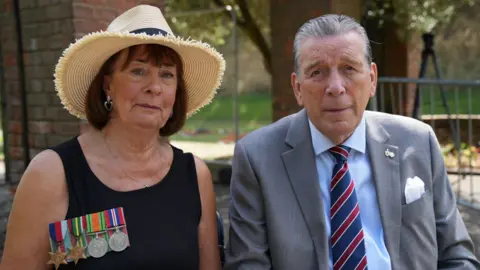 John Fairhall/BBC Gillian Powell sits with her husband Stephen on her left-hand side. She is wearing a black dress with her father's military medals attached. She is also wearing a straw hat and a silver necklace and earrings, and she has dark brown hair. Stephen is wearing a grey suit jacket with a light blue shirt and dark blue and red striped tie. He has a white handkerchief in his breast pocket, and has greying hair which is combed backwards.