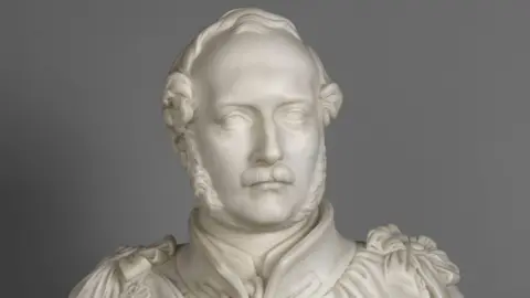 Royal Collection Trust The all white statue made of marble shows a young Prince Albert, wearing old Victorian era attire, with a small moustache and a beard which runs down each cheek and under his chin.