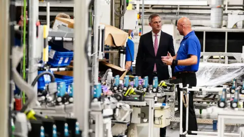 PA Media A man in a black suit and pink tie talking to another man, in a blue t-shirt, in a car factory, surrounded by machines with components.
