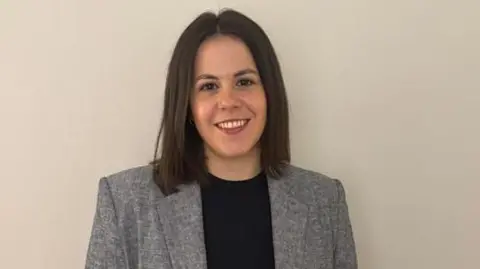 Silvia Prades A woman with brown hair wearing a grey jacket and a black top smiles in front of a plain wall.