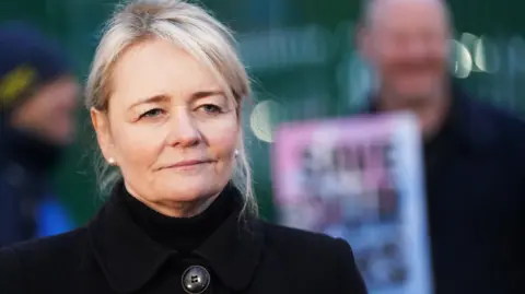 PA Media A woman with blonde hair, tied back, wears a black winter coat, buttoned up, and white pearl ear studs. She is standing on a picket line. Man can be seen holding placards in the background, which is out of focus.
