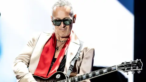 Getty Images Vivian Campbell is on stage performing. He's playing a guitar. He's wearing sunglasses and a silver jacket with a red scarf and necklaces. White and blue is shining behind him from a large screen.