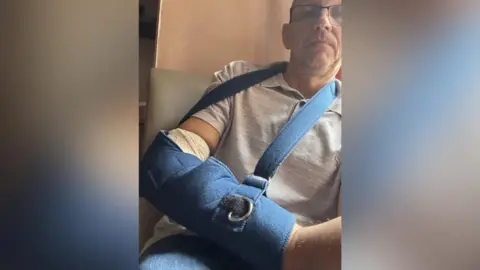 Handout A middle-aged man sits in a chair with his arm bandaged and in a blue sling. 